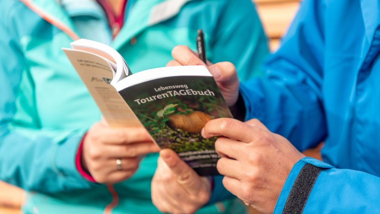 Zwei Personen in blauen Jacken halten ein TourenTAGEbuch und schreiben darin.