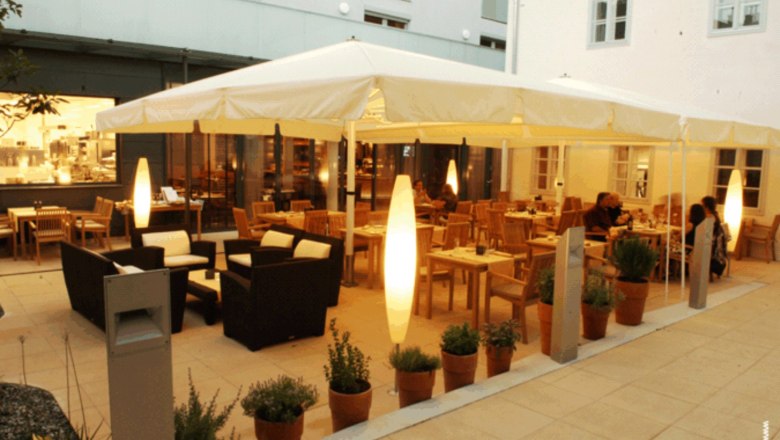 Ein beleuchteter Gastgarten mit Tischen, Stühlen und Pflanzen in Töpfen, umgeben von Gebäuden.