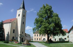 Kirche mit Turm und Uhr in einem Dorfplatz mit Bäumen und Gebäuden.