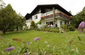 Ein zweistöckiges weißes Haus mit Balkon, umgeben von einem grünen Garten mit Blumen und Bäumen.