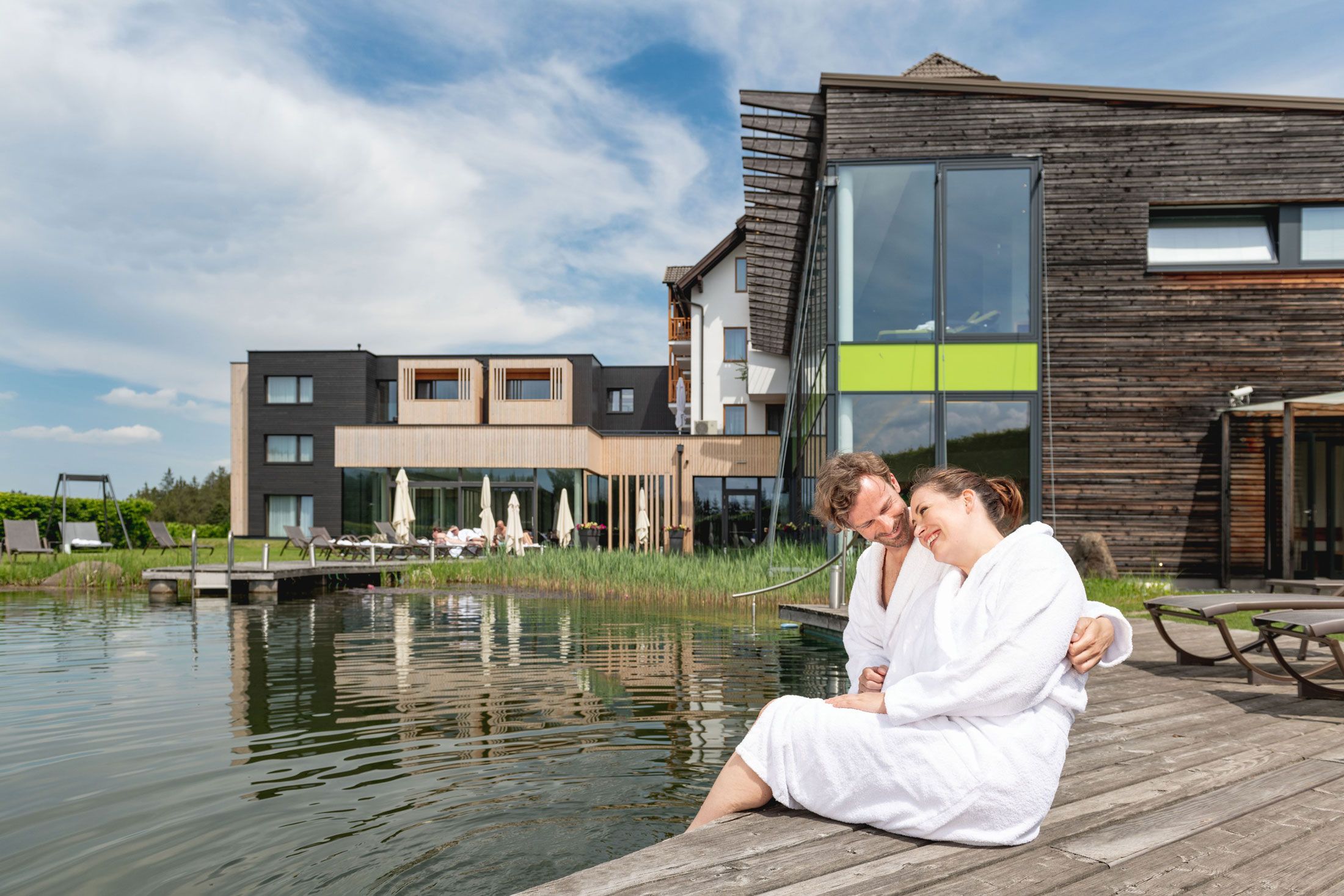 Ein Paar in weißen Bademänteln sitzt auf einem Holzsteg an einem Natur-Schwimmbiotop vor einem modernen Gebäude.
