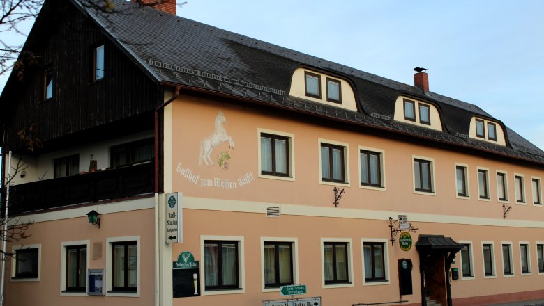 Gasthof „Zum weißen Rössel“, © Gasthof „Zum weißen Rössel“ Außenansicht des Gasthofs 'Zum weißen Rössel' mit beige Fassade und Pferdemalerei.