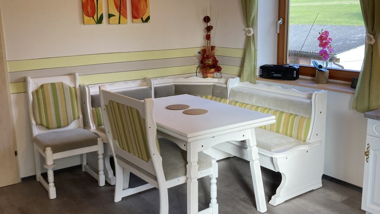 Gemütliche Essecke mit Holzmöbeln und grün-weiß gestreiften Polstern, Fenster mit Blick ins Grüne, Tulpenbild an der Wand.