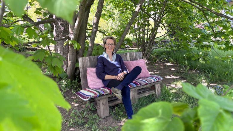 Eine Person sitzt auf einer Bank im Garten, umgeben von grünen Blättern und Bäumen.