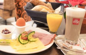 Ein Fr&uuml;hst&uuml;cksteller mit K&auml;se, Schinken, Ei, Marmelade, Butter, Br&ouml;tchen, Orangensaft und einem Latte Macchiato.