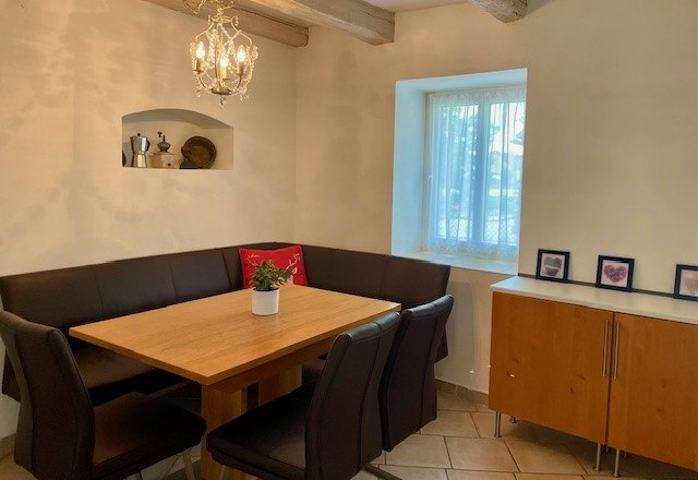 Ein gemütlicher Essbereich mit Holztisch, schwarzen Stühlen und Eckbank. An der Wand hängt ein Kronleuchter, daneben ein Fenster und ein Sideboard.