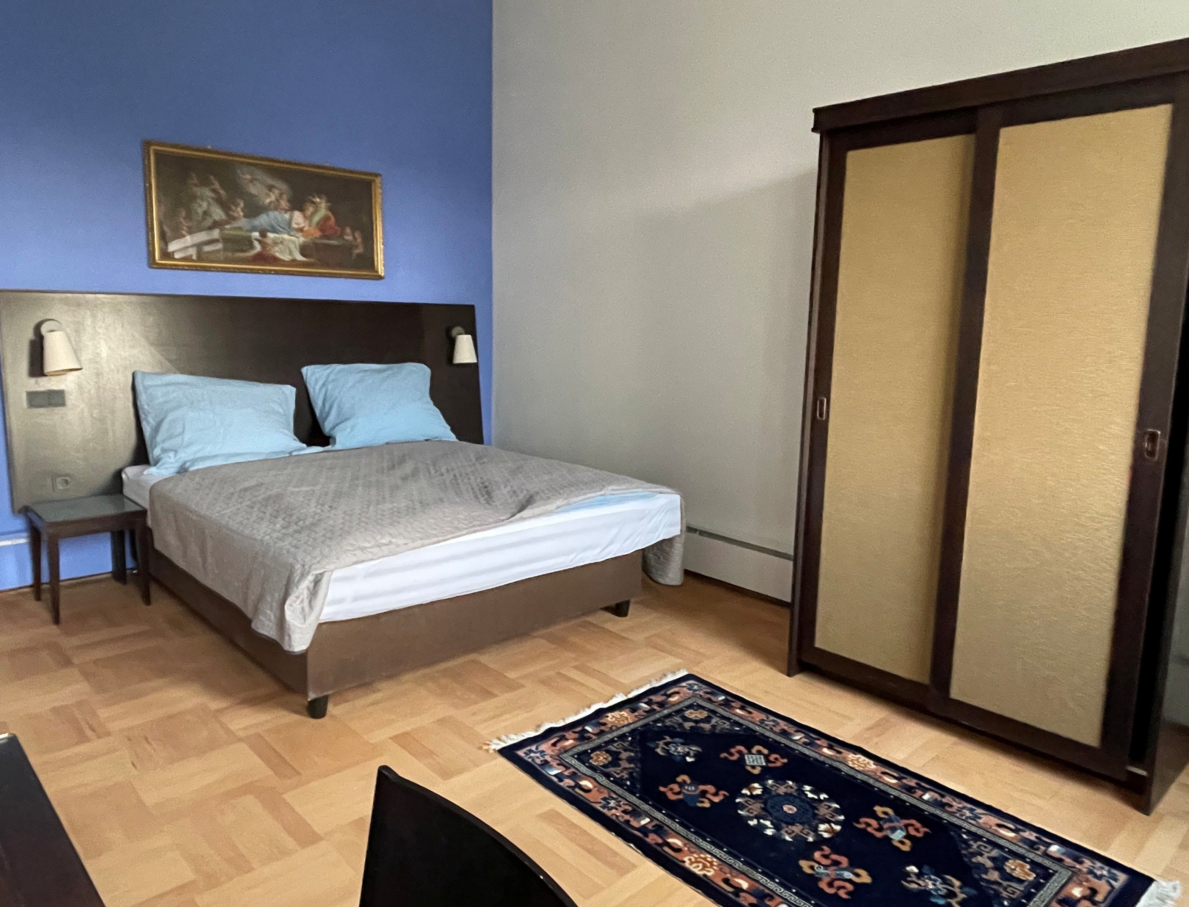 Ein Schlafzimmer mit blauer Wand, einem Doppelbett, einem Schrank und einem Teppich auf Holzboden.