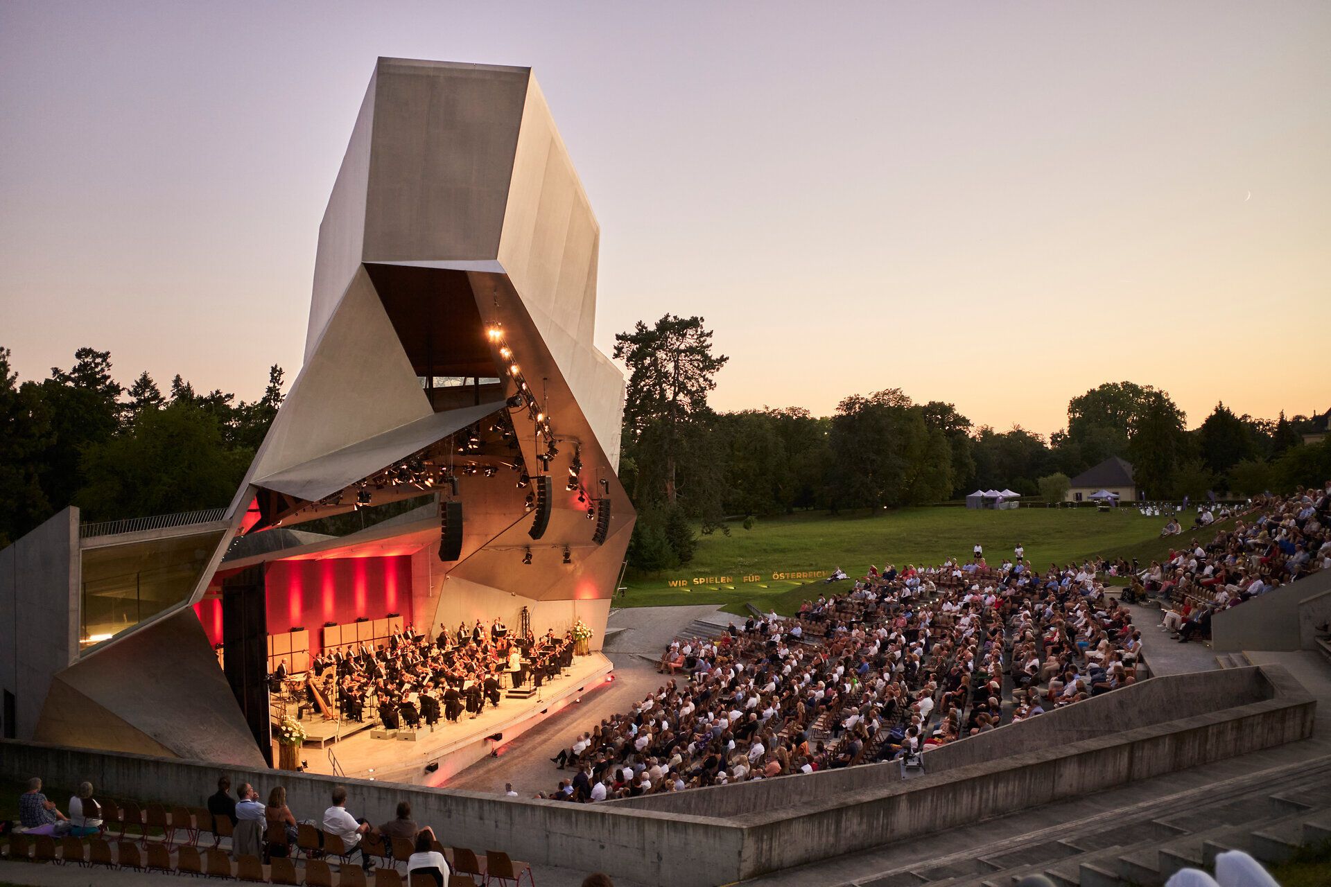 In der malerischen Kulisse von Grafenegg versammeln sich Musikliebhaber unter freiem Himmel, um die Klänge eines beeindruckenden Konzerts zu genießen. Die sanften Hügel und die umgebende Natur schaffen eine harmonische Atmosphäre, die den Sommerabend unvergesslich macht.