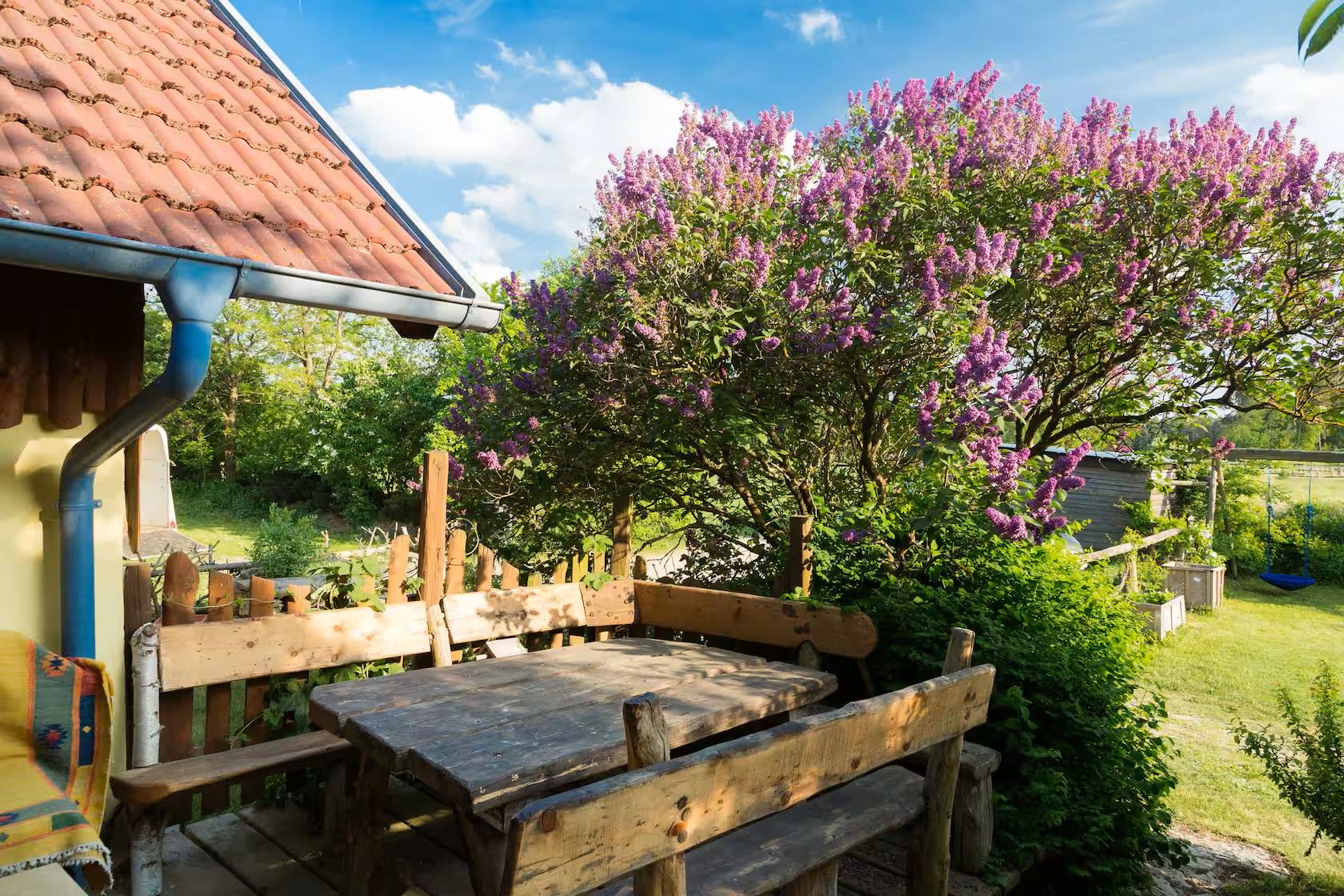 Terrasse mit Holztisch und Bank neben einem Haus mit rotem Ziegeldach; daneben blühender Flieder im Garten.