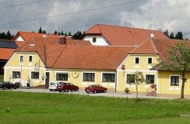 Gelbes Gebäude mit roten Dächern und mehreren Autos davor, umgeben von grüner Wiese und Bäumen.