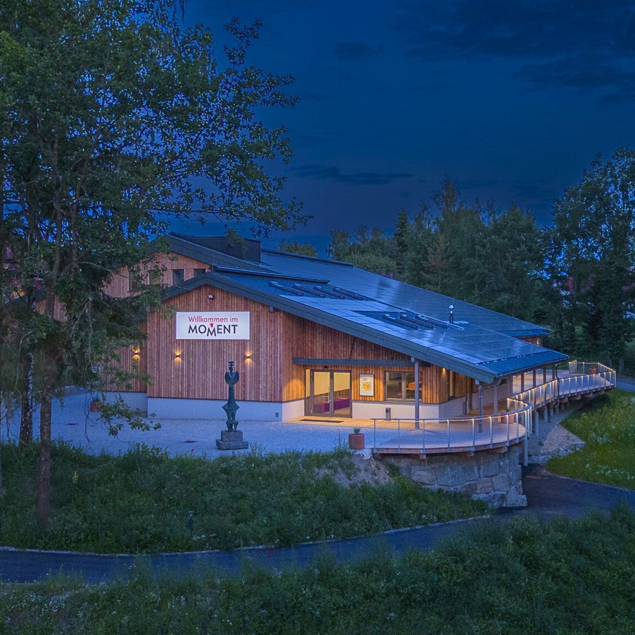 Ein modernes Gebäude aus Holz mit einem Schild "Willkommen im MOMENT" bei Nacht, umgeben von Bäumen.