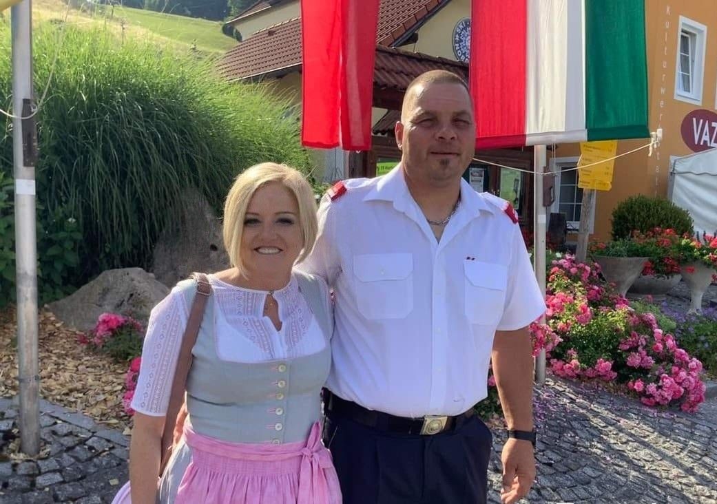 Ein Mann in Uniform und eine Frau in traditioneller Kleidung stehen lächelnd nebeneinander vor einem Gebäude mit Flaggen.