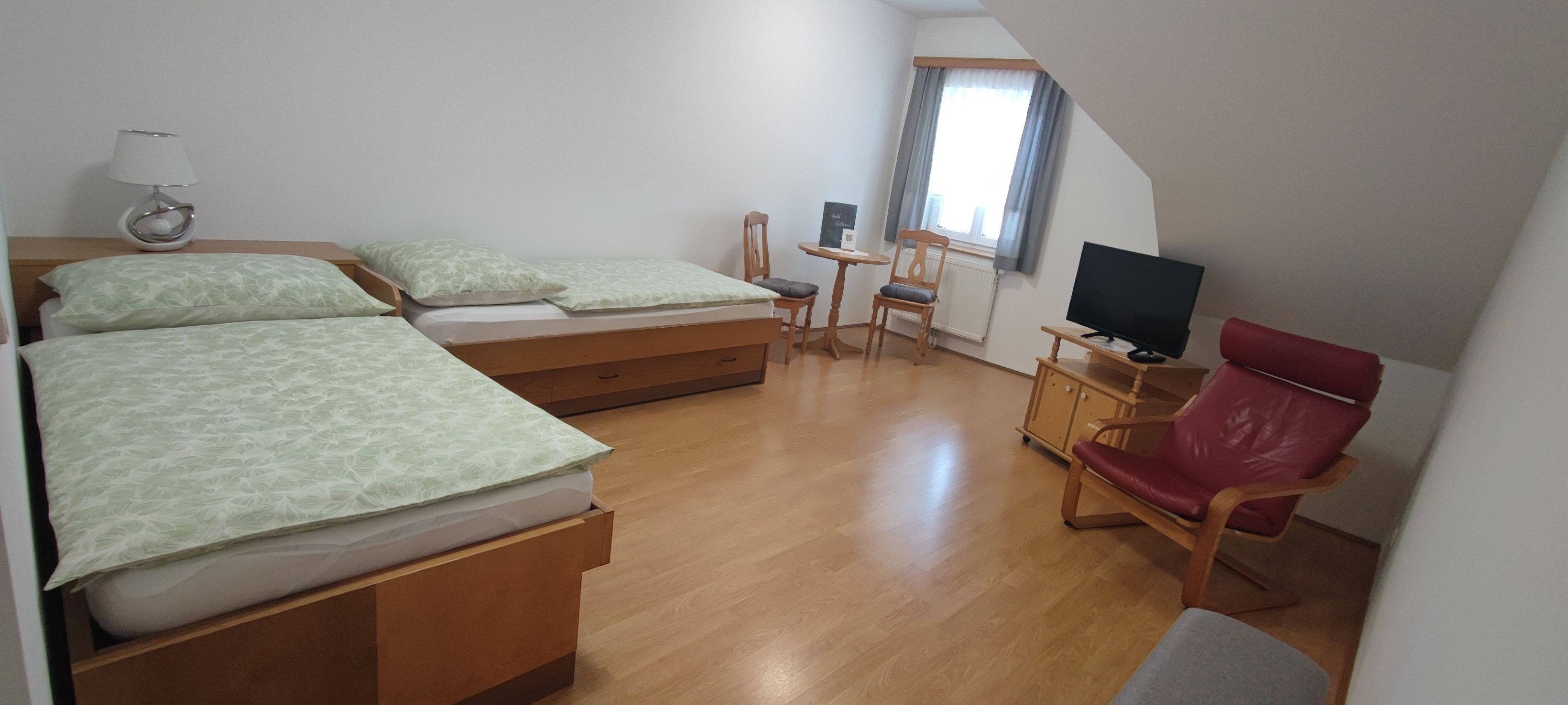 Ein einfach eingerichtetes Zimmer mit zwei Einzelbetten, einem kleinen Tisch mit Stühlen, einem Fernseher und einem roten Sessel.