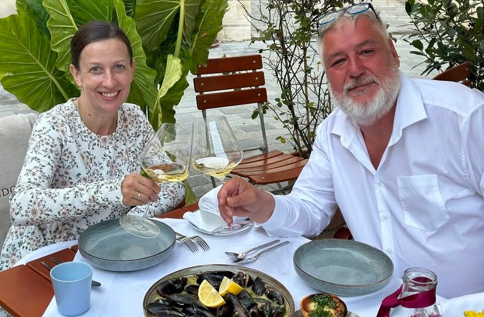 Ein Mann und eine Frau sitzen an einem Tisch im Freien und stoßen mit Weingläsern an. Vor ihnen steht ein Teller mit Muscheln.