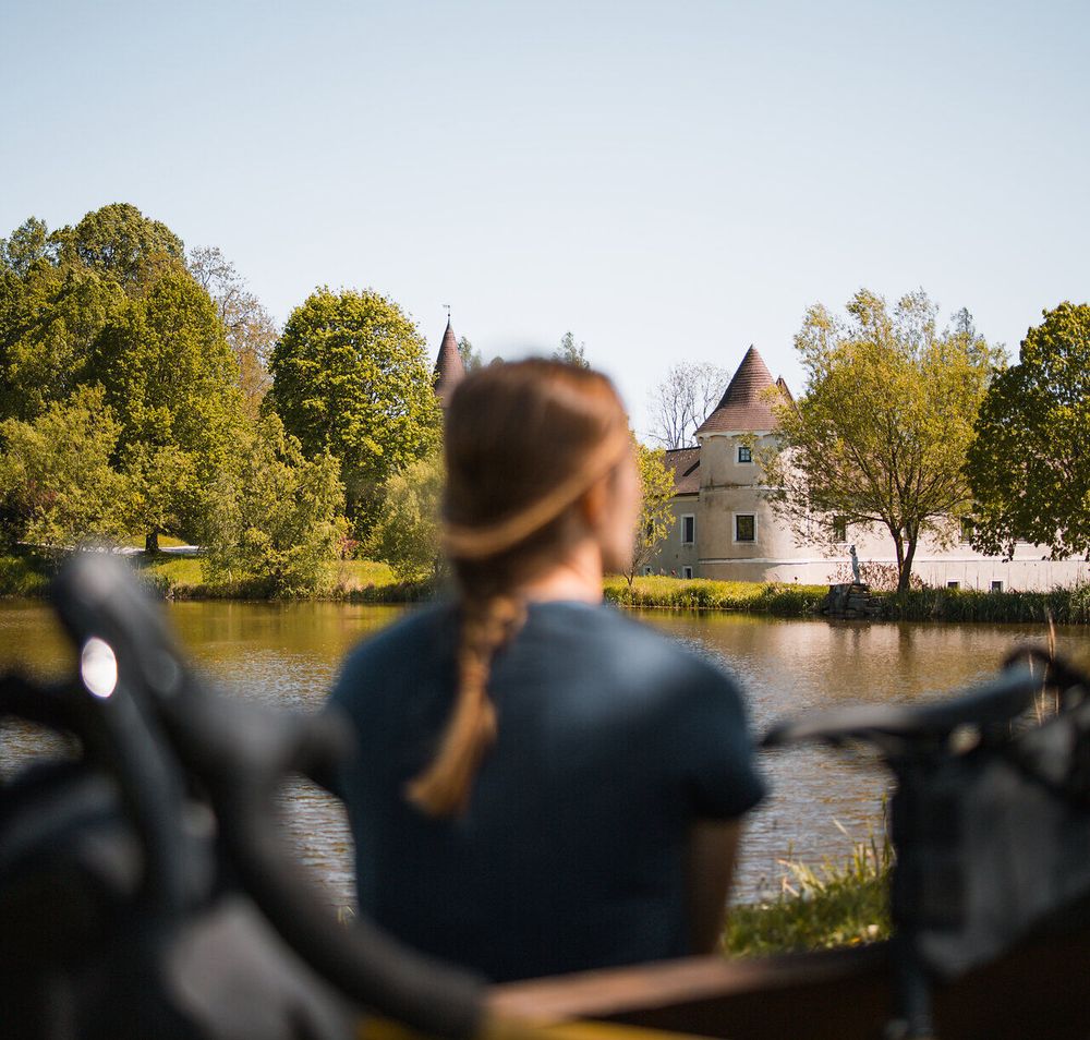 Ein sanfter Wind weht über das glitzernde Wasser, während die Bäume in sattem Grün erstrahlen. Die malerische Kulisse des Schlosses lädt Radfahrer ein, eine wohlverdiente Pause einzulegen und die Schönheit der Natur zu genießen.