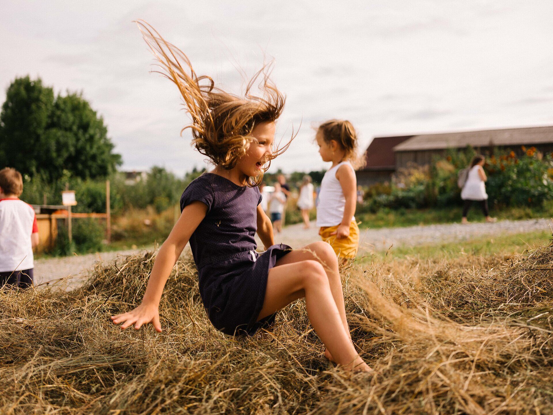 Kinder lachen und spielen fröhlich auf einem Haufen von Heu, während die sanfte Brise ihre Haare wehen lässt. Die warme Sonne taucht die Szene in ein goldenes Licht und schafft eine unbeschwerte Atmosphäre voller Lebensfreude und Abenteuer. Ein perfekter Tag, um die Natur und die Freude am Spiel zu genießen.