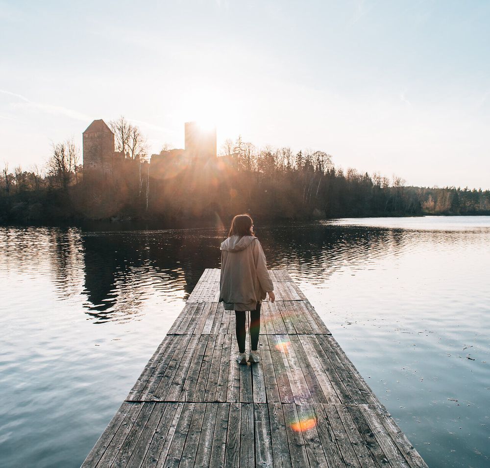 Ein sanfter Sonnenuntergang taucht die Landschaft in warmes Licht, während eine Person auf einem Holzsteg dem ruhigen Wasser entgegengeht. Die Stille der Natur und die sanften Wellen schaffen eine friedliche Atmosphäre, die zum Verweilen einlädt.