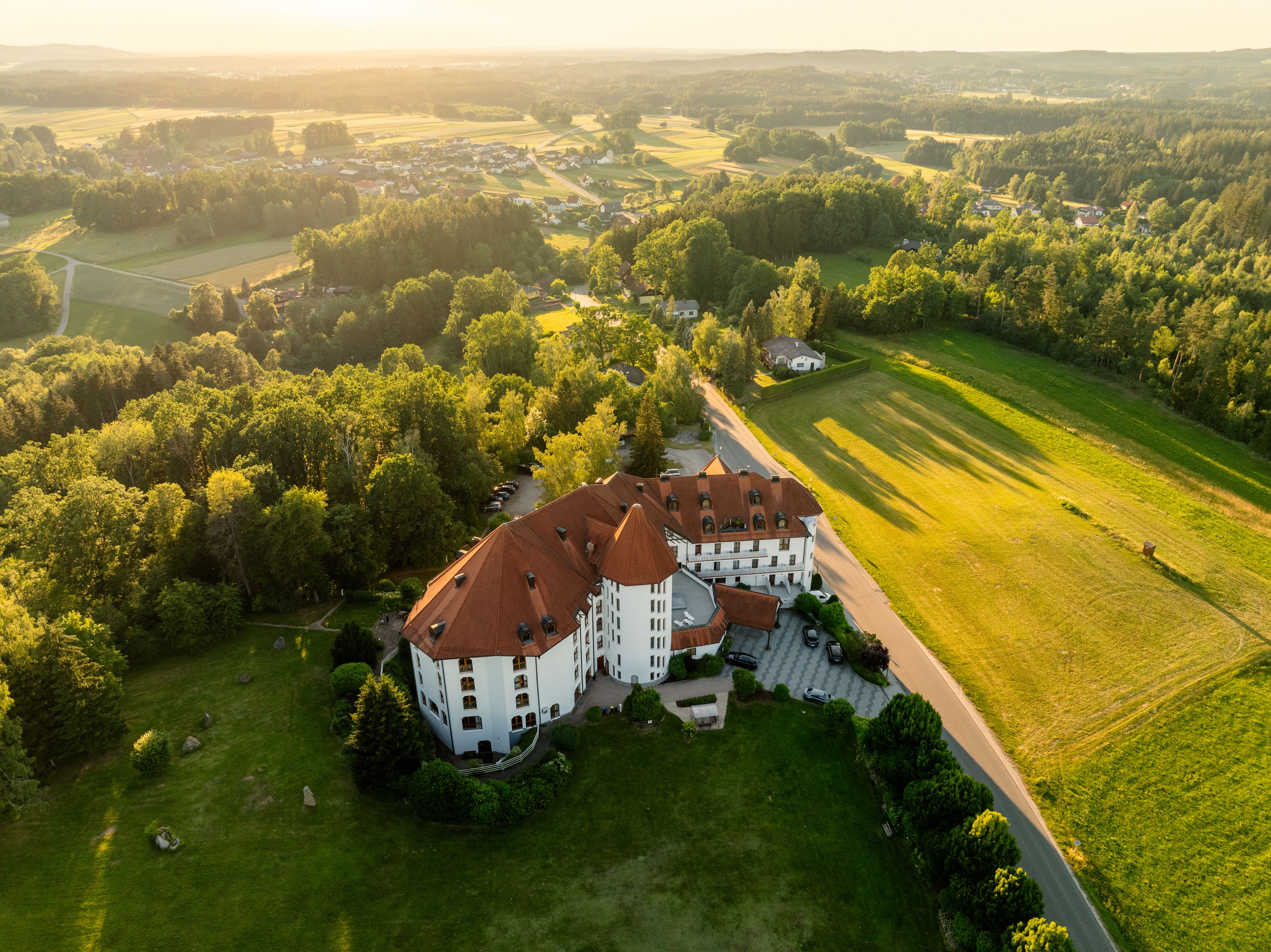 Luftaufnahme eines großen Gebäudes mit rotem Dach, umgeben von grünen Feldern und Wäldern bei Sonnenuntergang.
