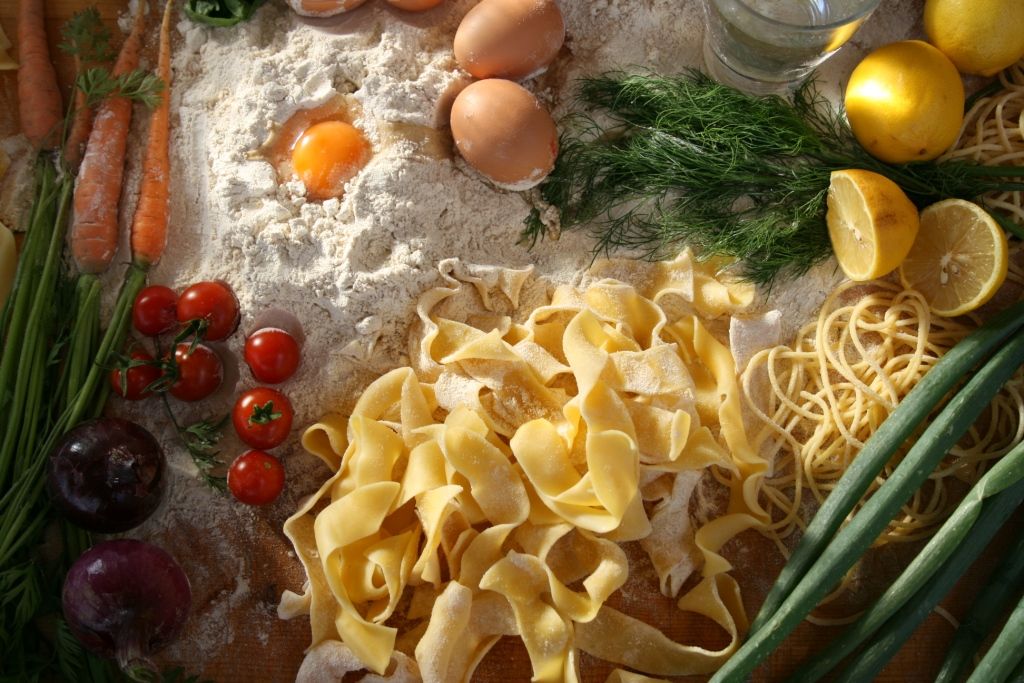 Zutaten für die Nudelzubereitung, darunter Eier, Mehl, Tomaten, Karotten, Zitronen und frische Kräuter.