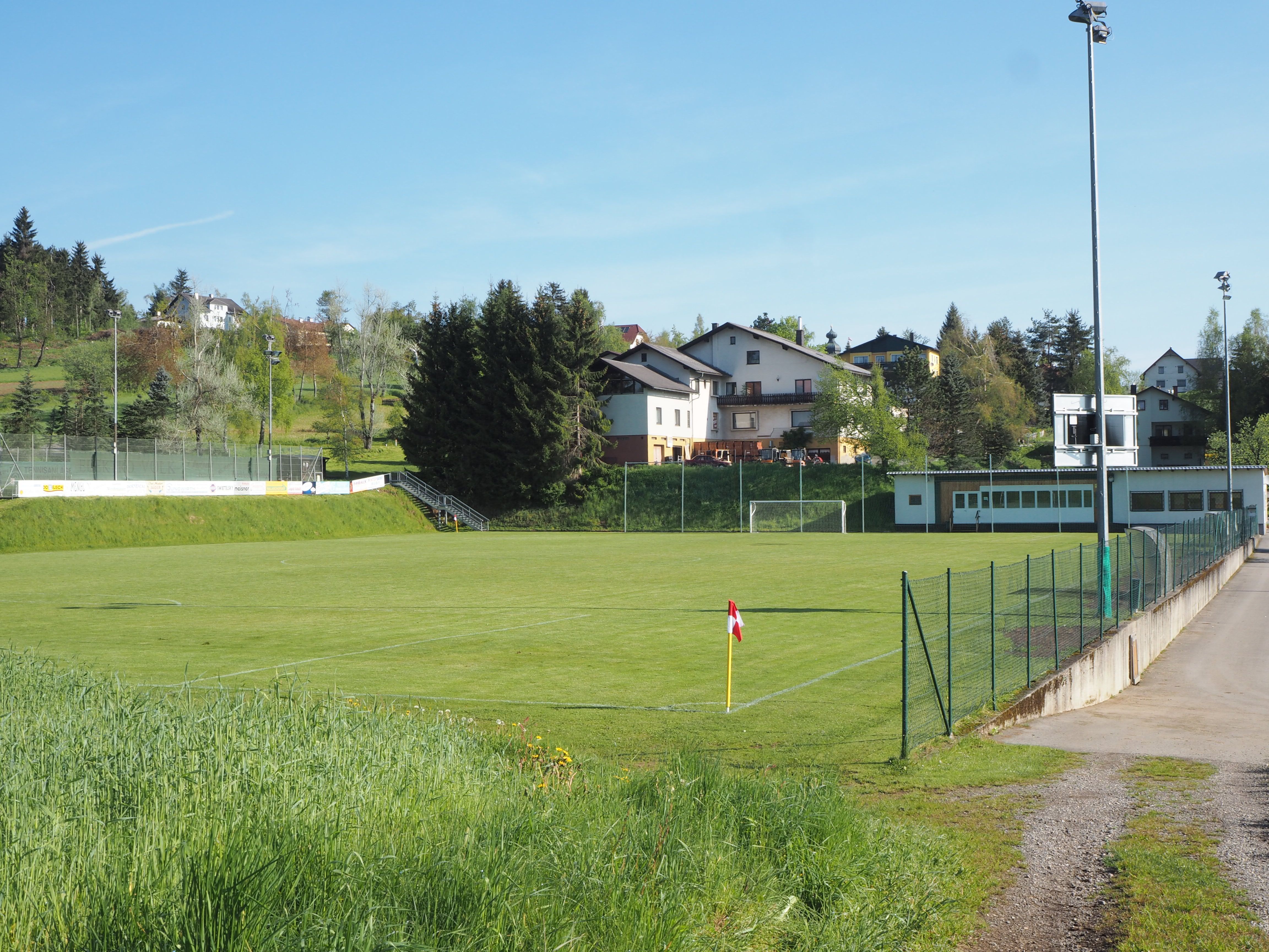 Ein leerer Fußballplatz mit umliegenden Gebäuden und Bäumen an einem sonnigen Tag.