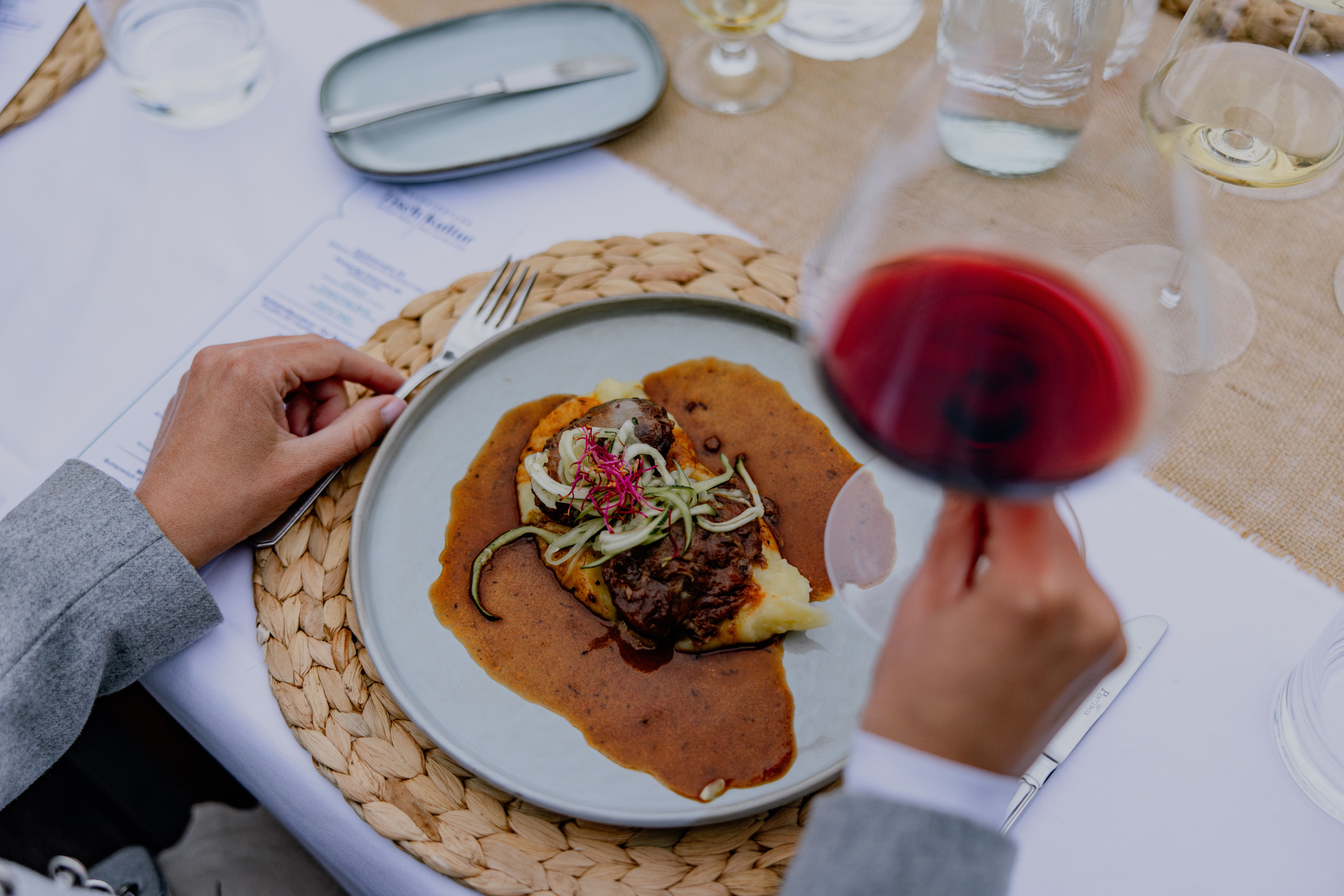 Person hält ein Glas Rotwein über einem Teller mit Fleischgericht und Soße.