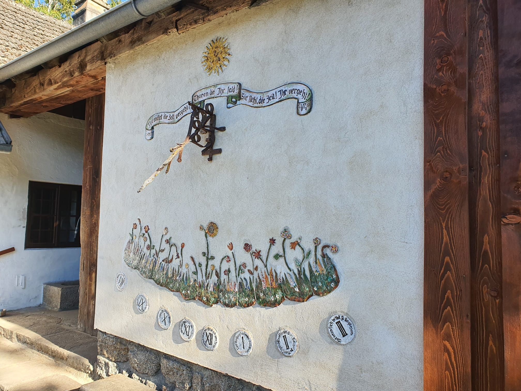 Sonnenuhr an einer Hauswand mit Blumenverzierung und Spruchband.