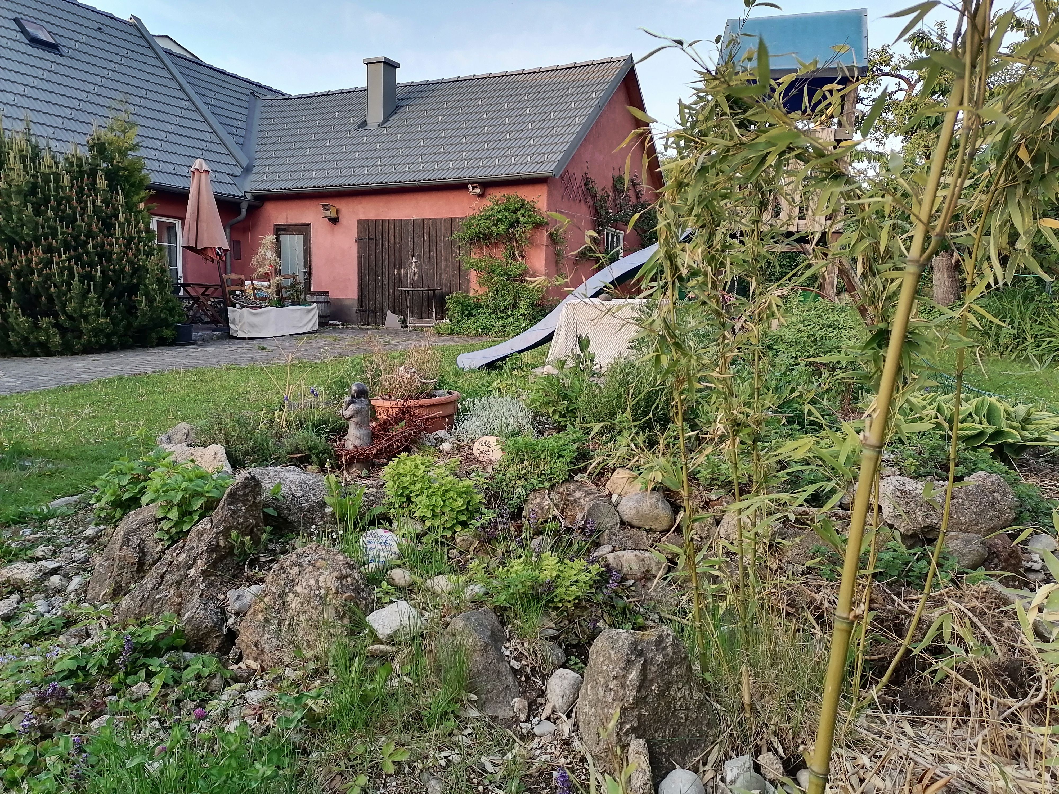 Ein Garten mit Steingarten, Bambus und einem roten Haus im Hintergrund. Eine kleine Statue steht zwischen den Pflanzen.