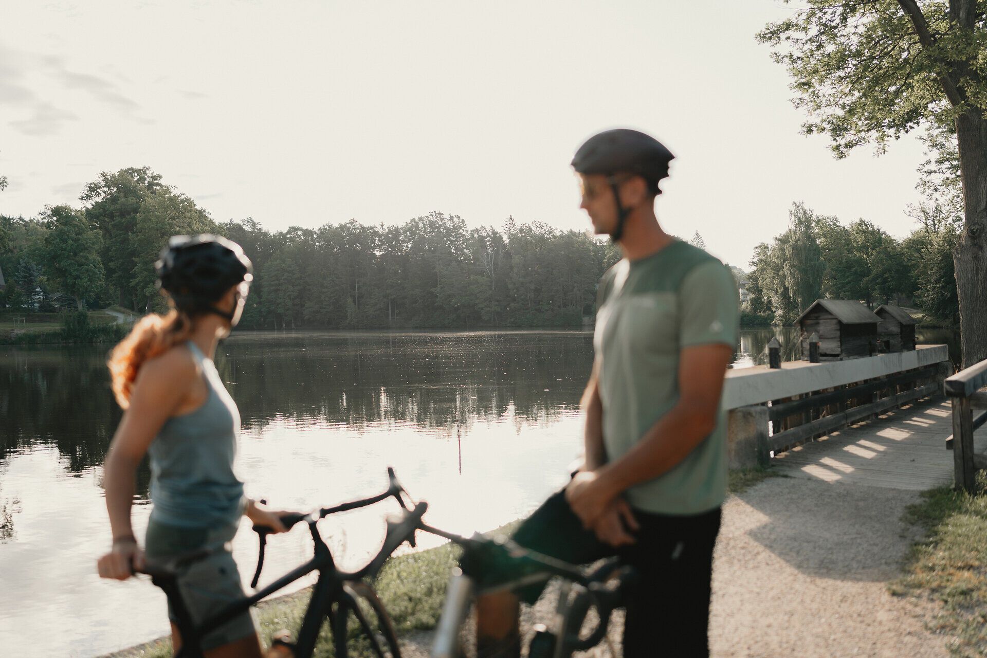 Zwei Radfahrer:innen mit Helmen stehen mit ihren Fahrrädern am Ufer eines ruhigen Teichs, umgeben von Wald.