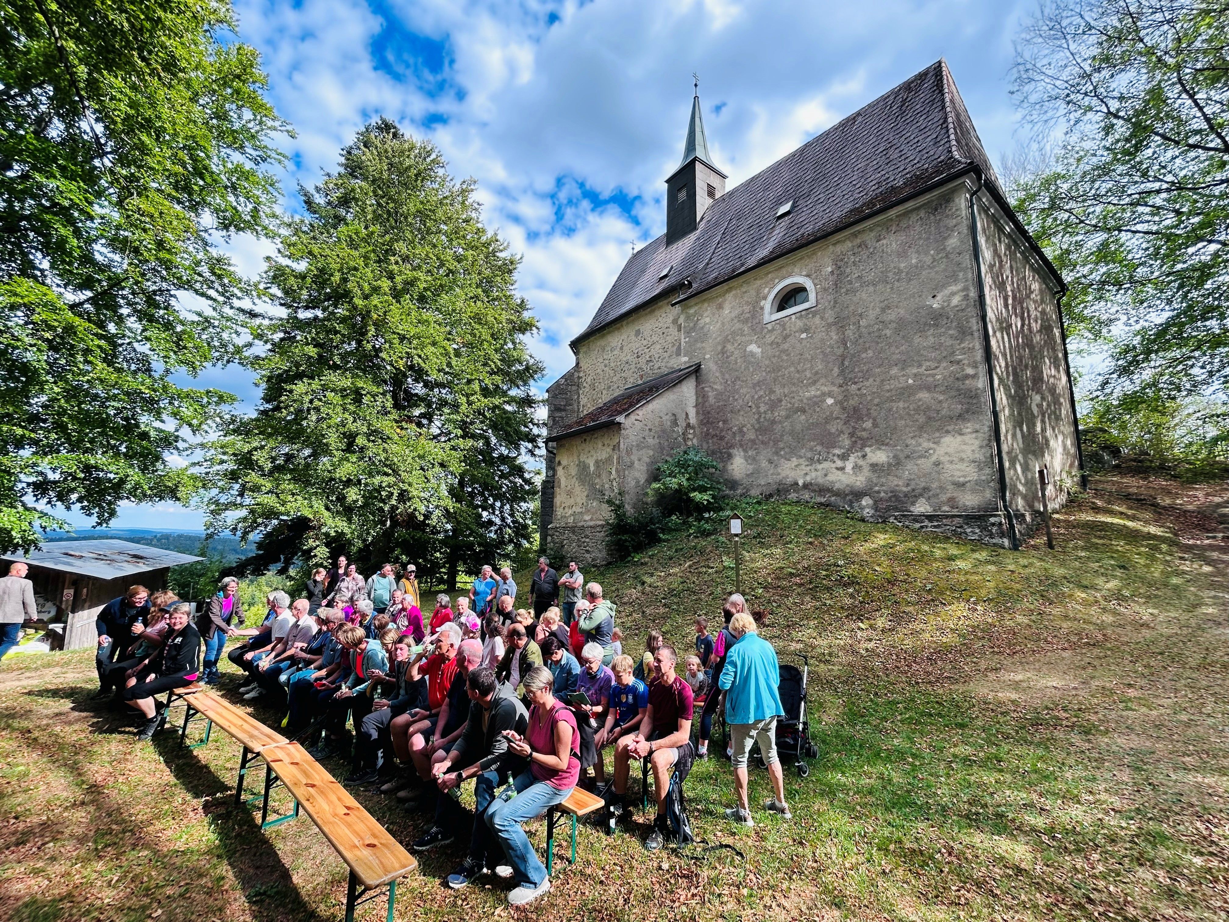 Eine Gruppe aus Menschen nimmt Teil an der Segnung des Marterls am Johannesberg als Teil der Station Gipfel der Visionen am Pilgerweg Bernhardiweg. Sie sitzen auf Heurigenbänken. Im Hintergrund sieht man die Johannesbergkirche. 
