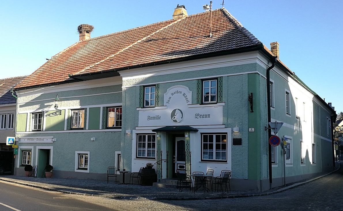 Ein traditionelles Gasthaus mit grüner Fassade und dem Namen 'Zum weißen Rössl'. Vor dem Gebäude stehen Tische und Stühle.