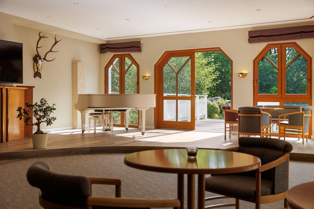 Ein heller Raum mit einem weißen Flügel, Holzmöbeln und großen Fenstern, die auf eine Terrasse mit Blick ins Grüne führen.