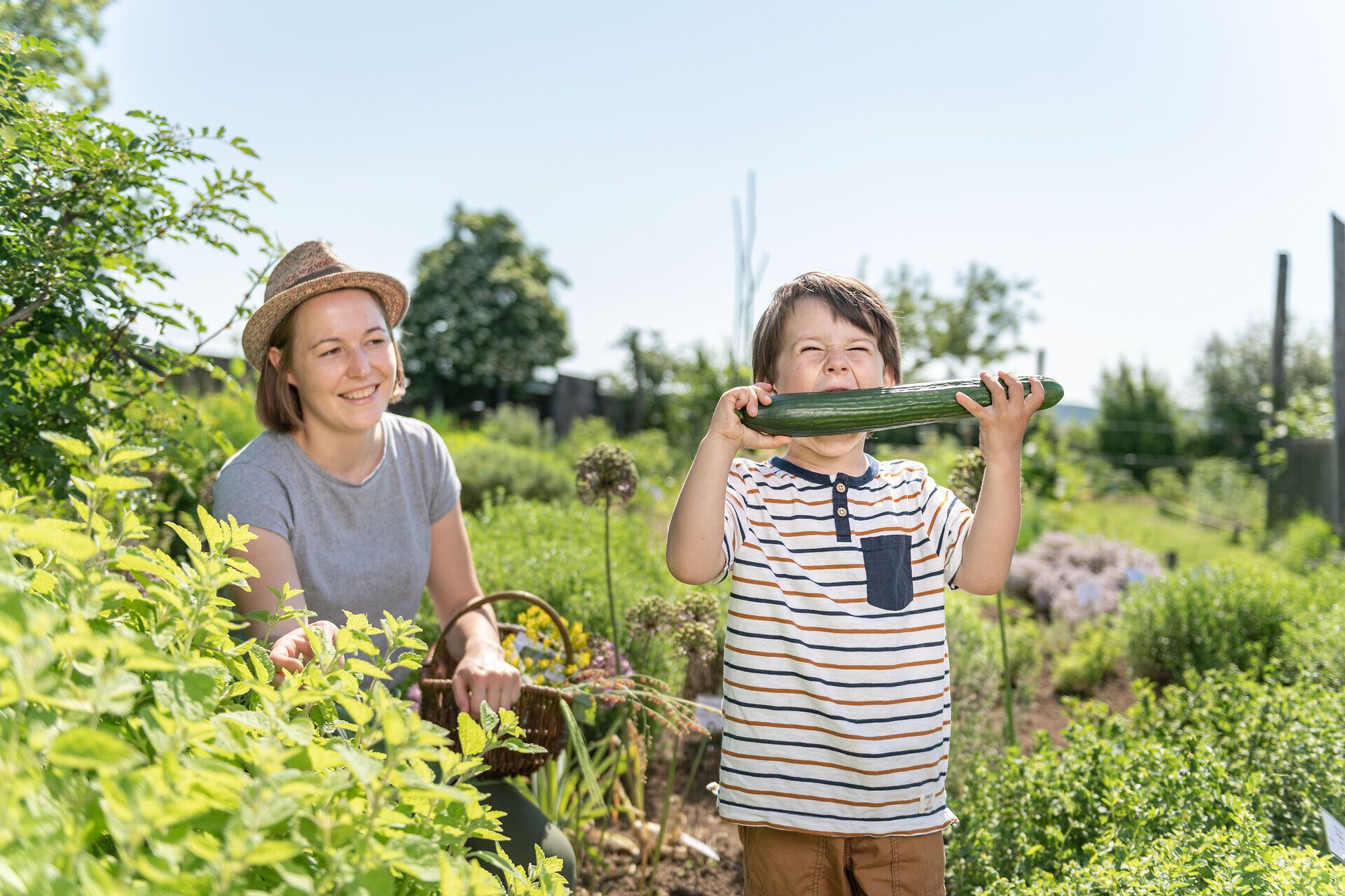 In einem blühenden Garten voller frischer Kräuter und Gemüse genießen ein Kind und eine Frau die Freude des Erntens. Die strahlende Sonne und die üppige Natur schaffen eine einladende Atmosphäre, die zum Verweilen und Entdecken einlädt.