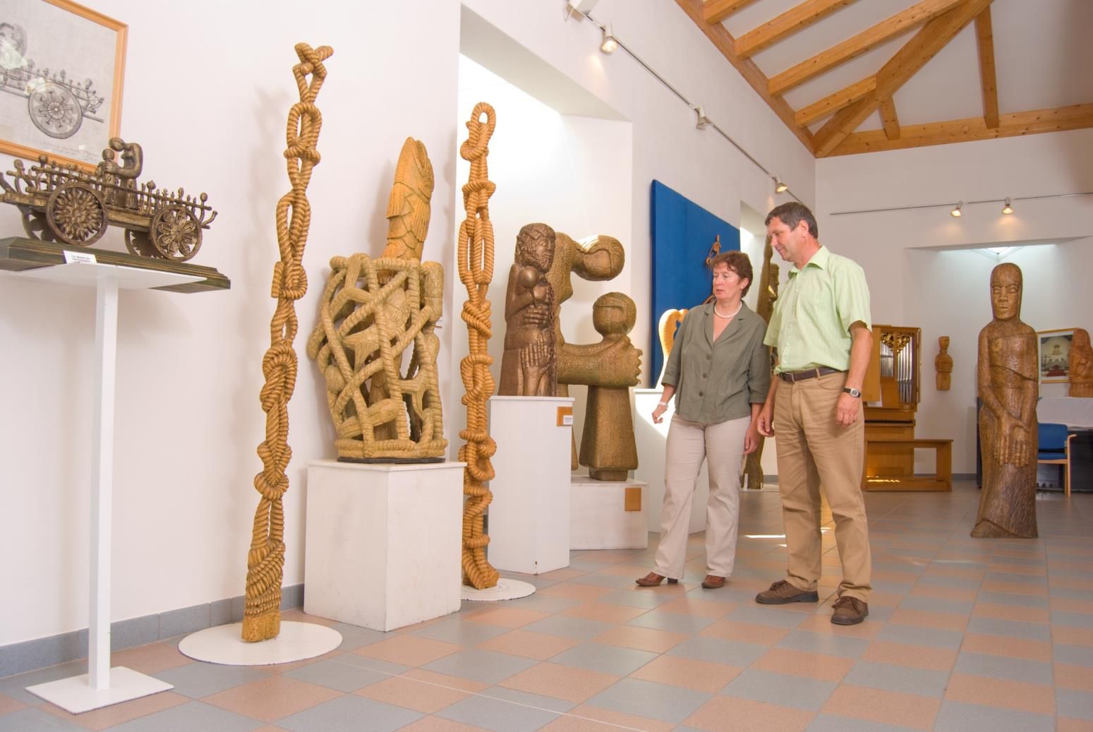 Zwei Personen betrachten Holzskulpturen in einem Ausstellungsraum mit Holzbalkendecke.