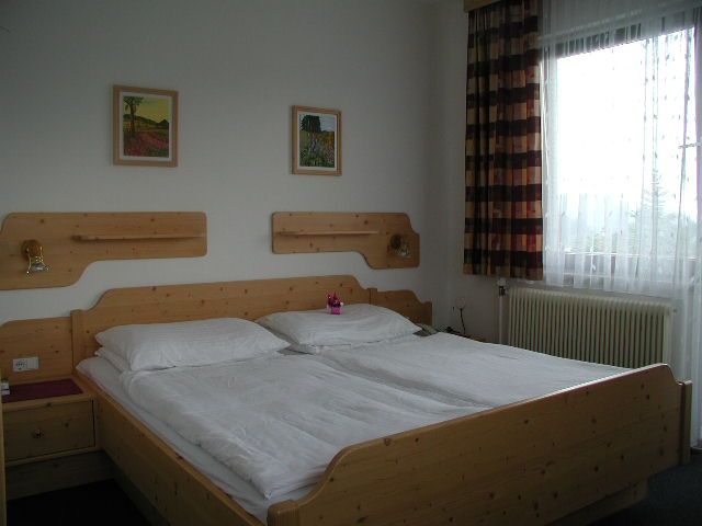 Ein einfach eingerichtetes Hotelzimmer mit einem Doppelbett aus Holz, zwei Bildern an der Wand und einem Fenster mit Vorhängen.