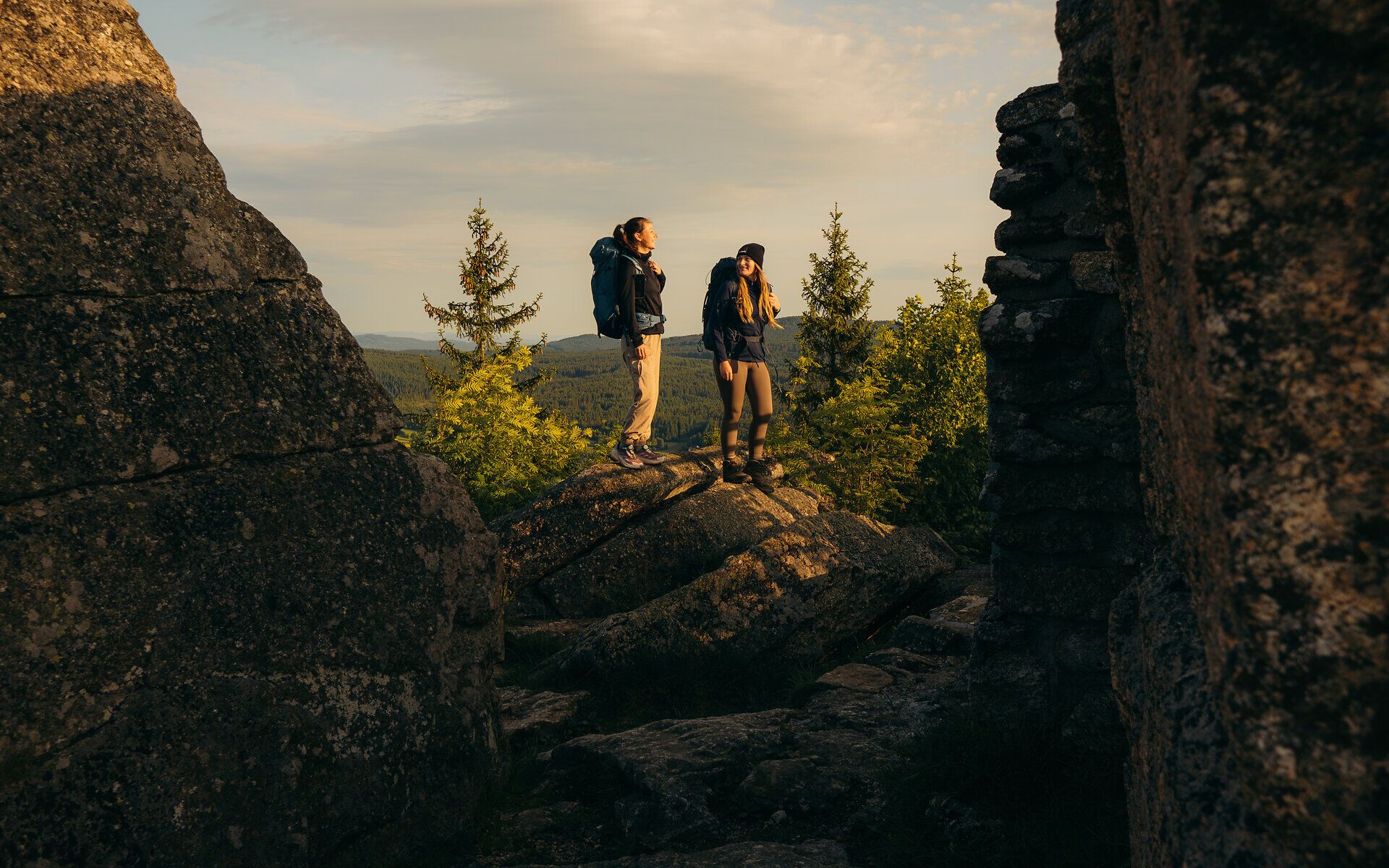 Zwei Wanderer stehen zwischen imposanten Felsformationen und genießen den weiten Blick auf die Waldlandschaft im goldenen Morgenlicht im Waldviertel.