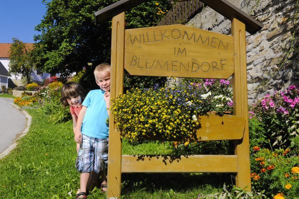 Zwei Kinder stehen neben einem Holzschild mit der Aufschrift 'Willkommen im Blumendorf', umgeben von bunten Blumen.
