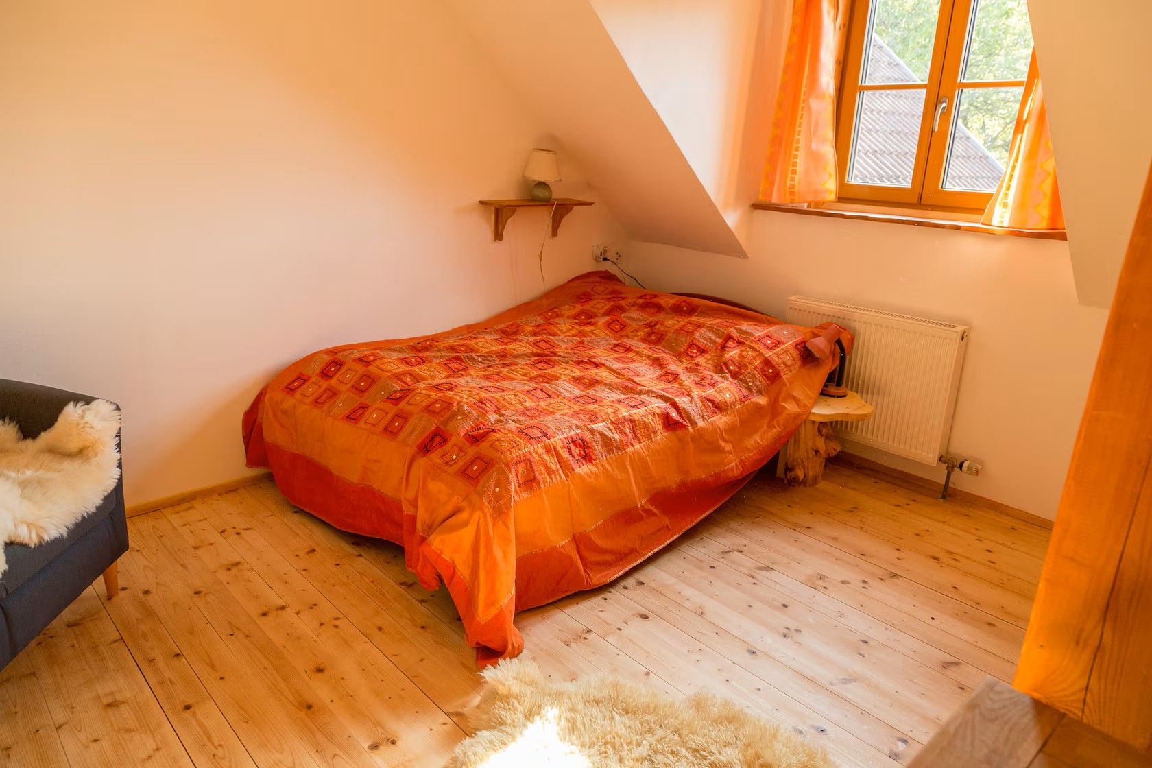 Gemütliches Schlafzimmer mit orangefarbener Bettdecke, Dachschräge, Fenster mit Vorhängen, Holzfußboden und kleiner Lampe auf dem Wandregal.