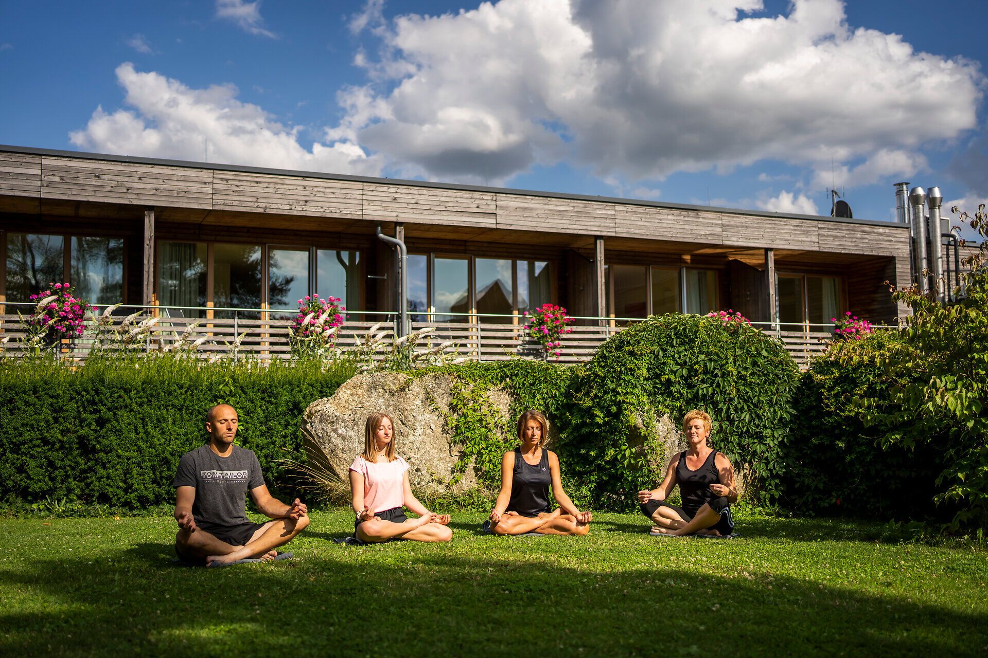 Inmitten einer malerischen Landschaft meditieren vier Personen im Grünen, umgeben von üppigem Gras und blühenden Pflanzen. Die sanften Wolken am Himmel und die harmonische Atmosphäre laden dazu ein, die innere Ruhe zu finden und die Schönheit der Natur zu genießen.
