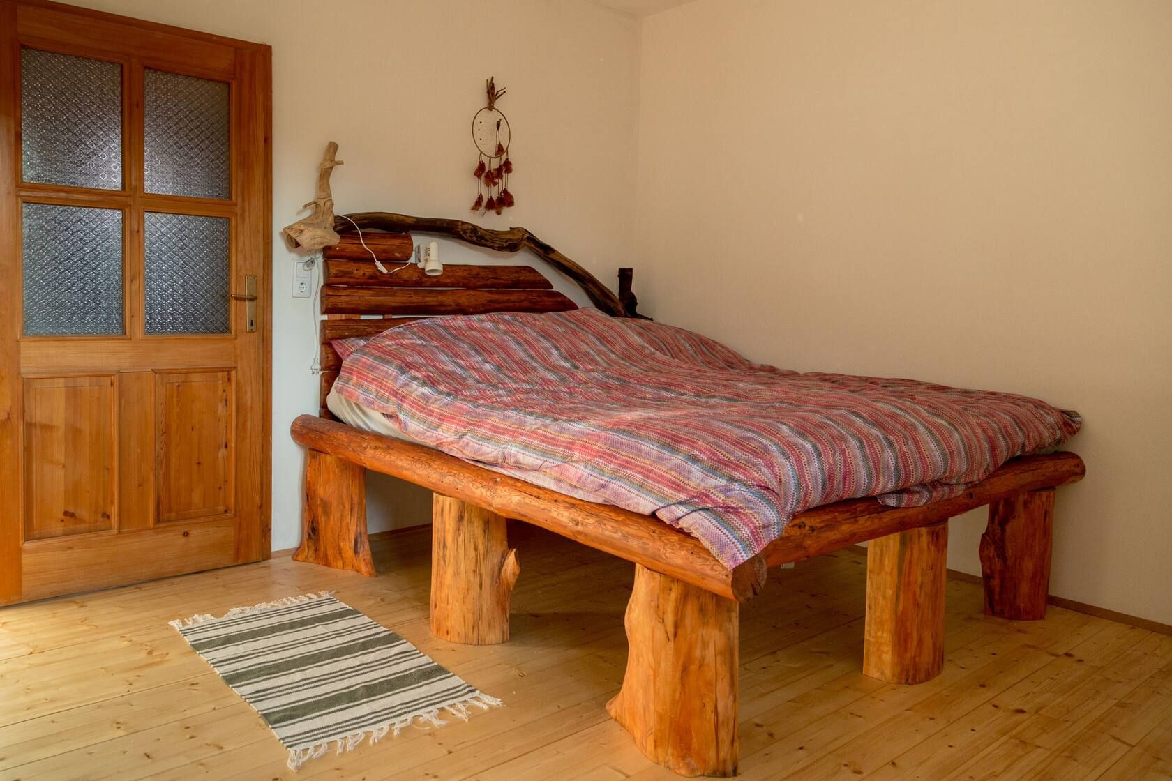 Rustikales Schlafzimmer mit Holzbett, gemusterter Decke, Holztür und kleinem Teppich auf hellem Holzboden.