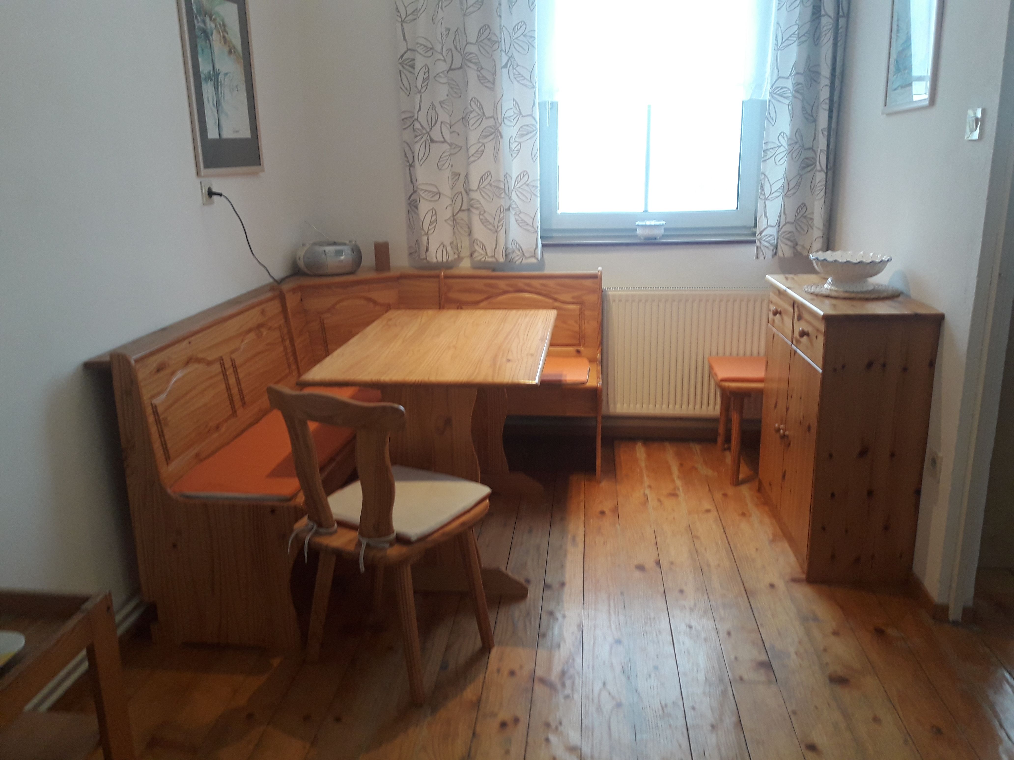 Gemütliche Küche mit Holztisch, Eckbank, Stühlen, Kommode und hellem Fenster mit gemusterten Vorhängen.