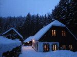 Verschneites Haus im Wald bei Dämmerung mit beleuchteten Fenstern.