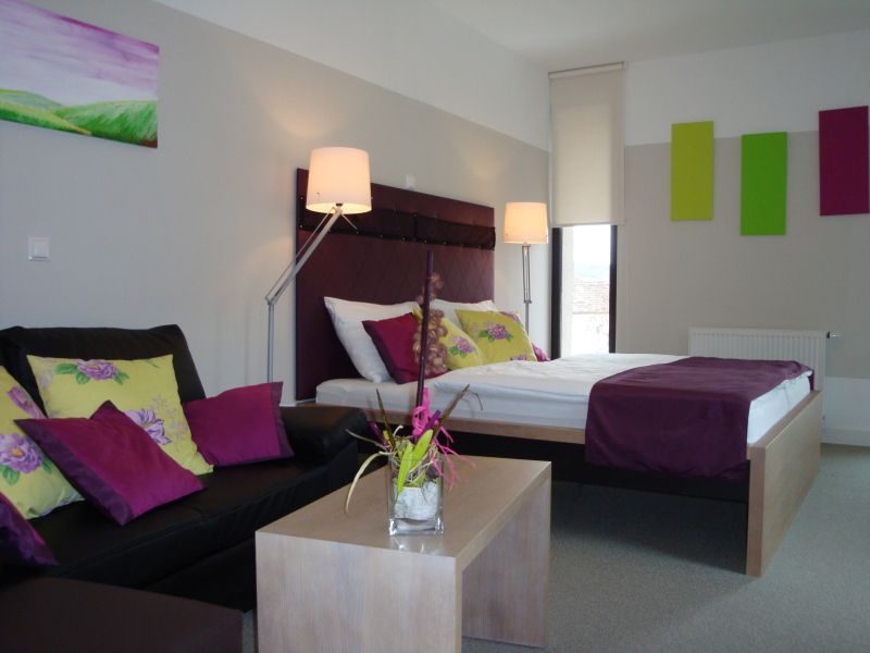 Modern eingerichtetes Hotelzimmer mit Doppelbett, Sofa und Dekoration in Lila- und Grüntönen.