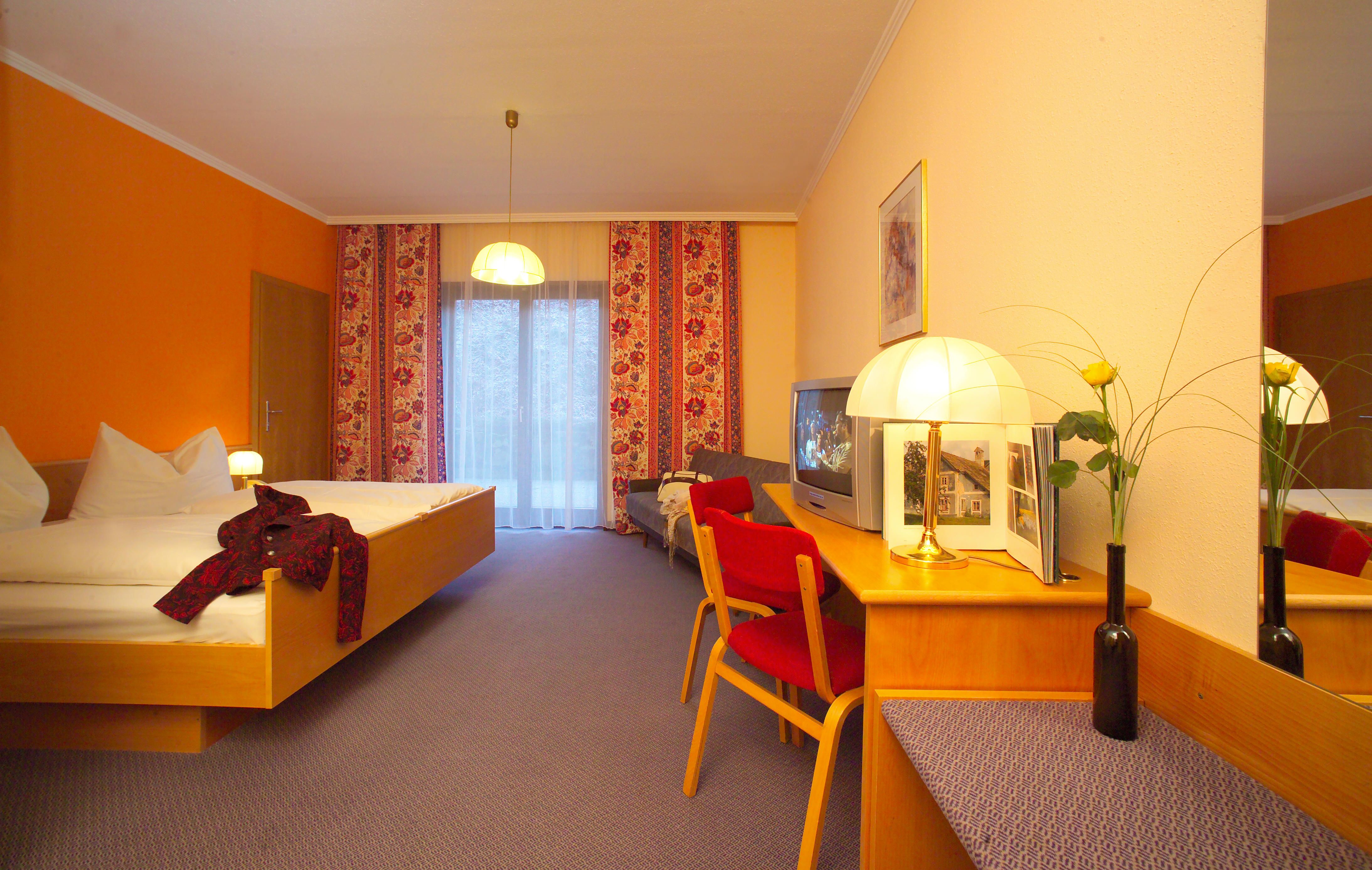 Hotelzimmer mit Doppelbett, rotem Stuhl, Fernseher und Lampe auf einem Tisch. Gelbe Wand und Vorhänge mit Blumenmuster.