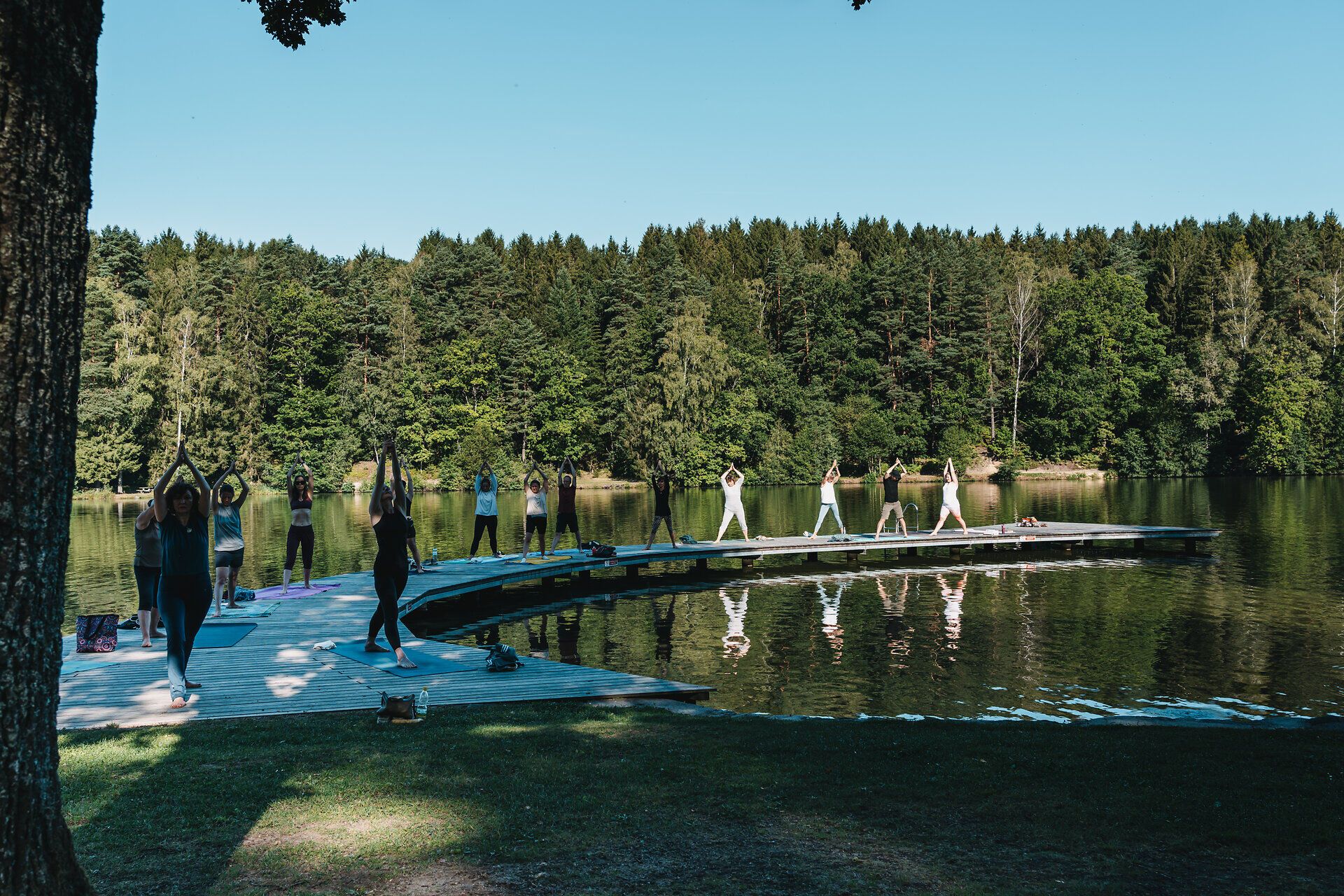 Inmitten der idyllischen Natur versammeln sich Menschen am Ufer eines ruhigen Sees, um Yoga zu praktizieren. Die sanften Bewegungen und die frische Luft schaffen eine harmonische Atmosphäre, die Körper und Geist belebt. Umgeben von üppigem Grün und dem sanften Plätschern des Wassers wird diese Erfahrung zu einem unvergesslichen Moment der Entspannung.
