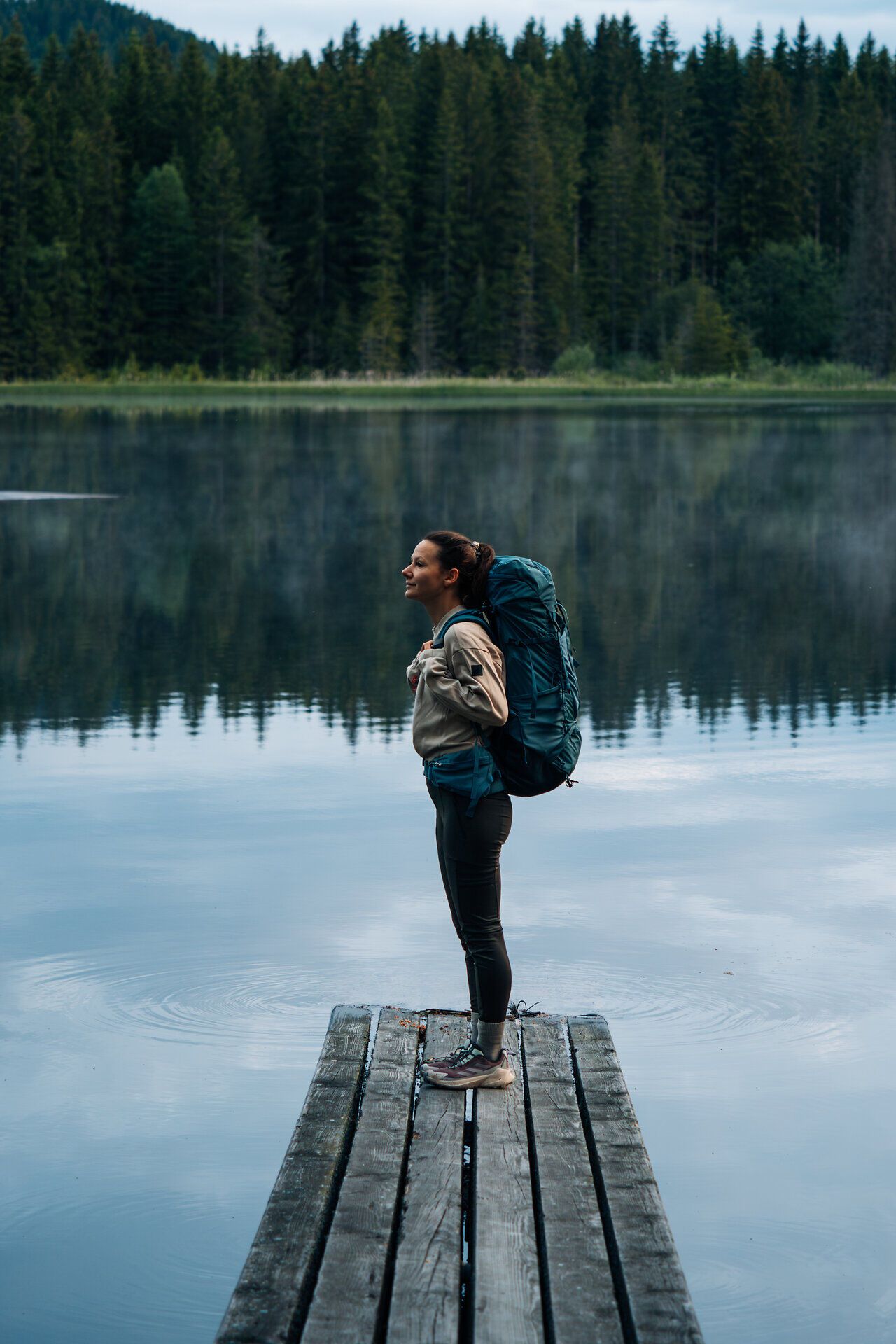 Frau steht auf einem Holzsteg am stillen Stierhübelteich, umgeben von dichter Waldlandschaft im Waldviertel, im Lichterspiel der Dämmerung.