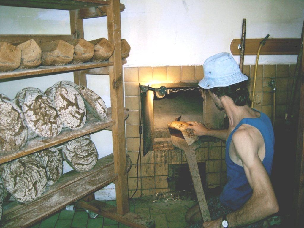 Ein Bäcker in einem blauen Tanktop und Hut nimmt Brot aus einem Ofen. Im Hintergrund sind Regale mit Brotlaiben zu sehen.