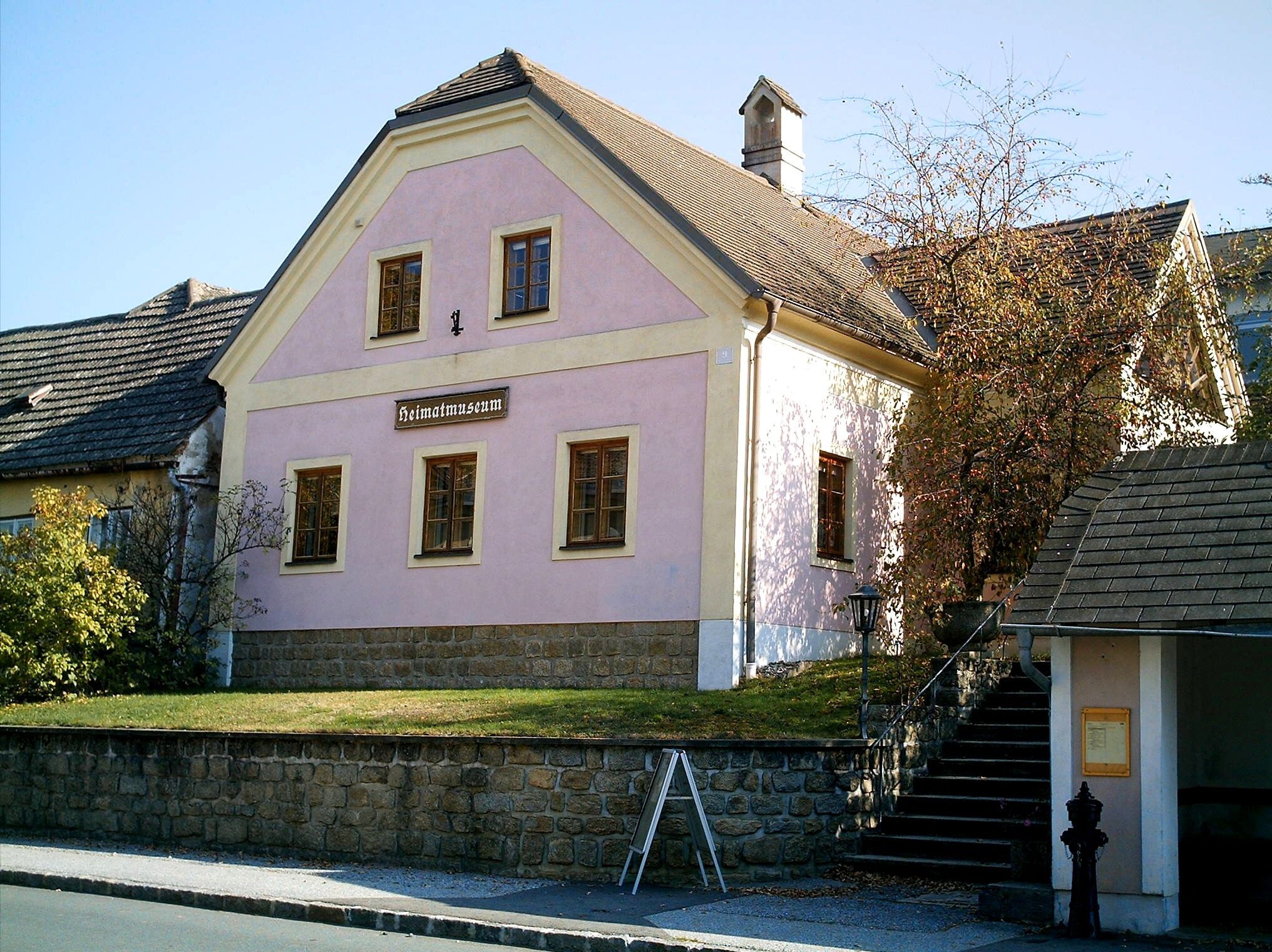 Ein rosa Gebäude mit der Aufschrift 'Heimatmuseum' in Kautzen, umgeben von Bäumen und einem gepflasterten Weg.