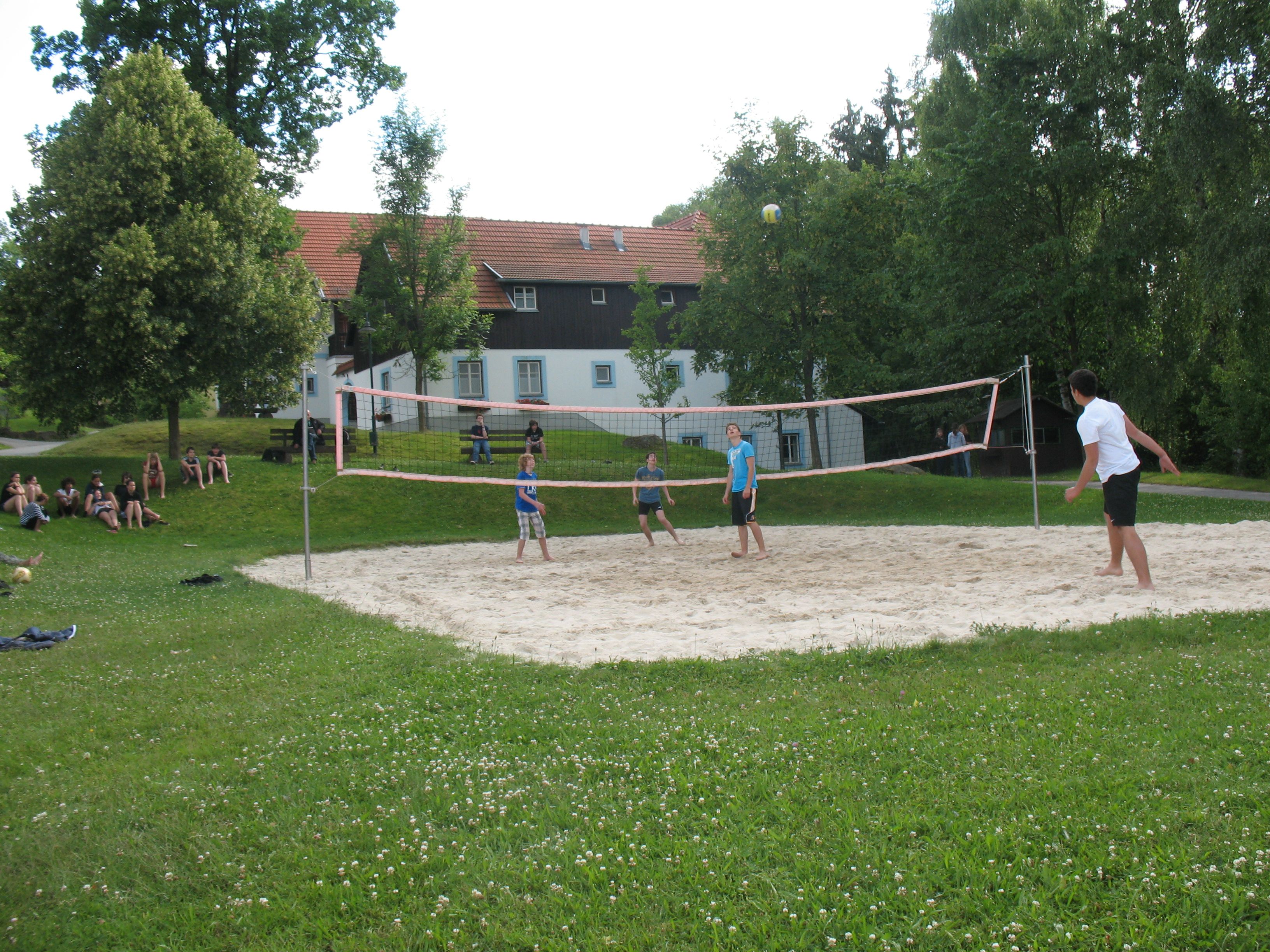 Menschen spielen Volleyball auf einem Sandplatz im Freien, umgeben von Bäumen und einem Gebäude im Hintergrund.