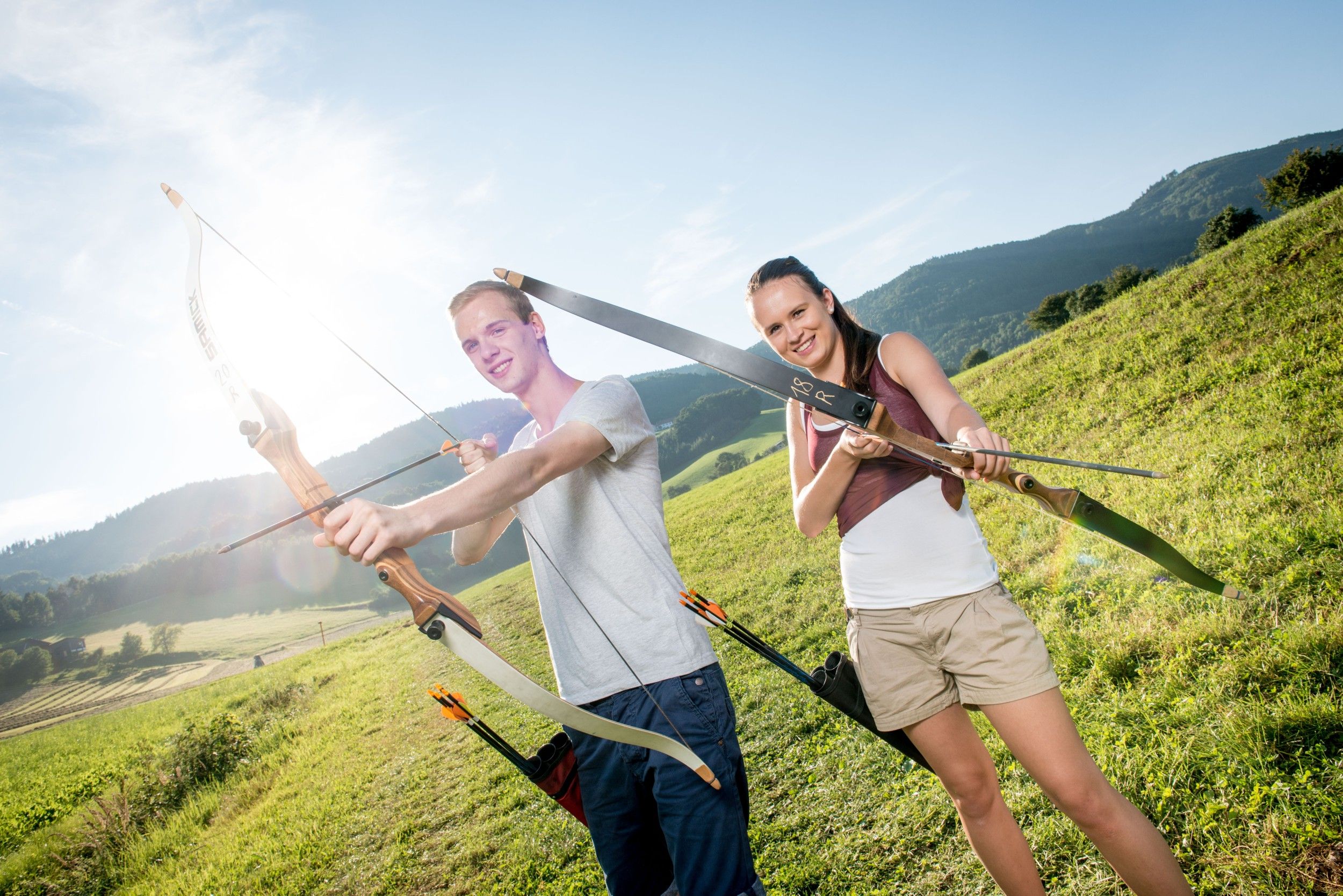 Zwei Personen beim Bogenschießen auf einer Wiese mit Bergen im Hintergrund.
