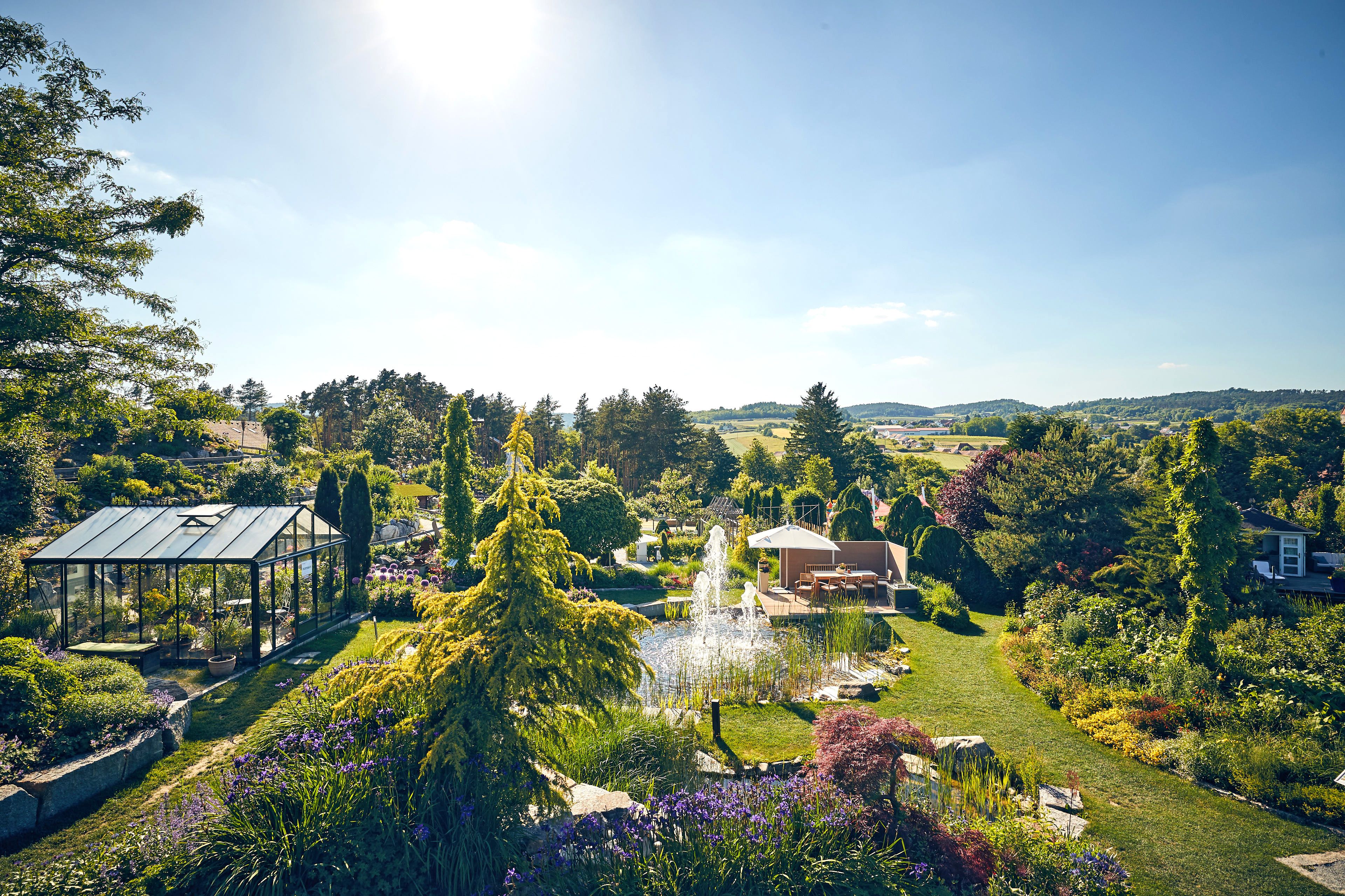 Ein Springbrunnen und ein Glashaus mitten in verschiedene Blumen und Bäumen mit Aussicht auf hügelige Landschaft.
